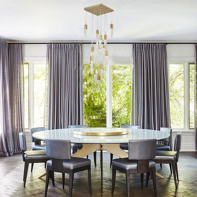 The 24-light gold cluster chandelier centered above a large round dining table in a room with floor-to-ceiling windows and grey drapery.